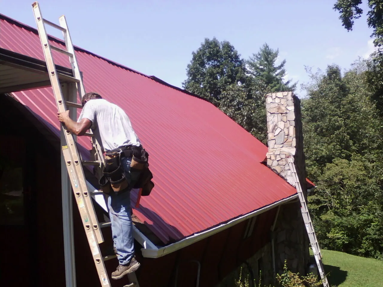 Roofer on a ladder beside a red metal roof and stone chimney — Soffit & Fascia Repair by Roofing Brothers in Klemme